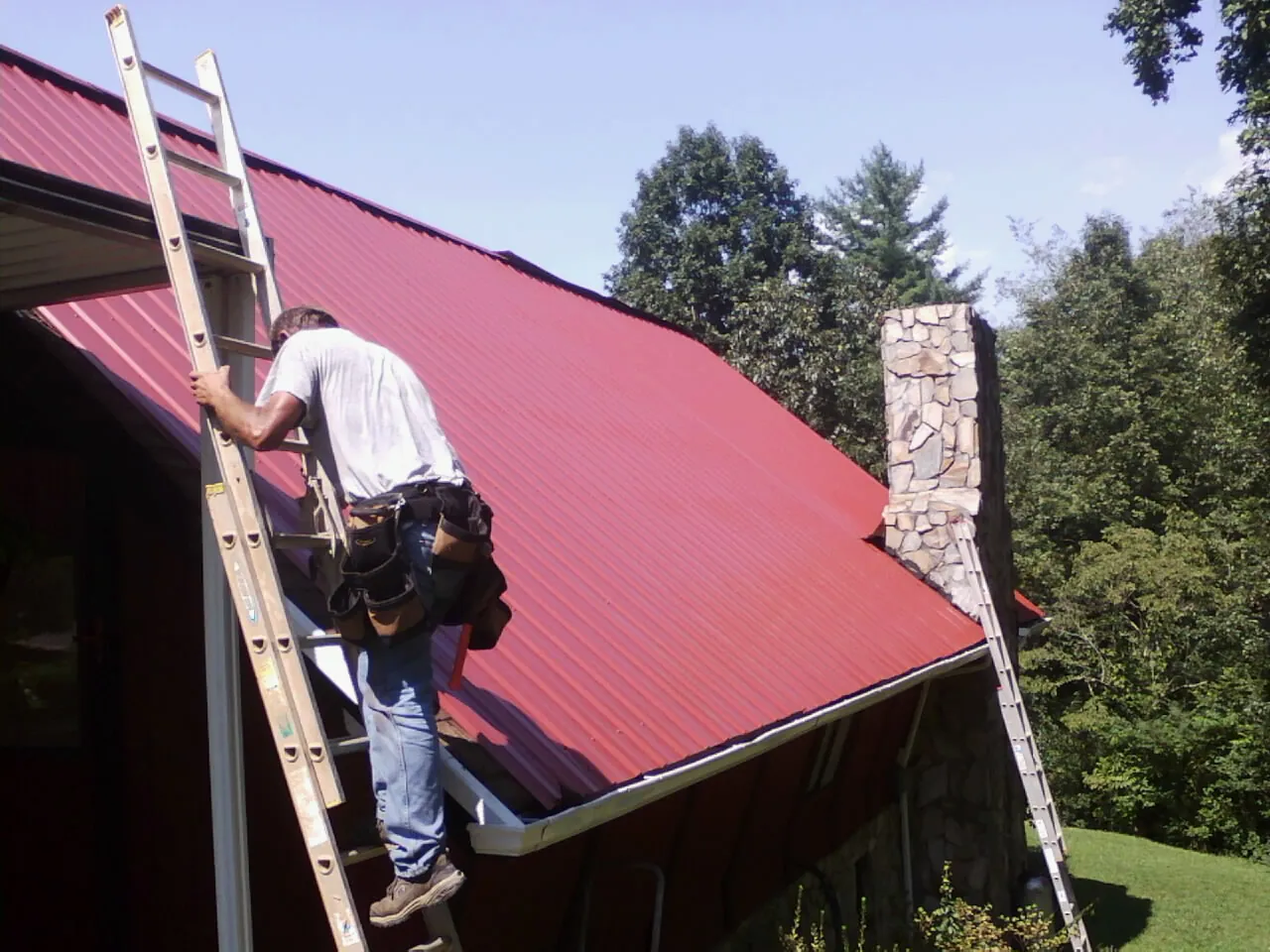 Roofer on a ladder beside a red metal roof and stone chimney — Soffit & Fascia Repair by Roofing Brothers in Woodside